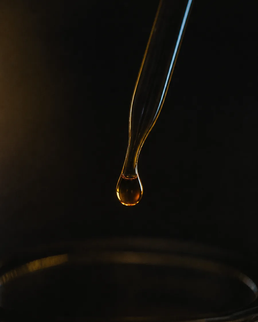 A single amber droplet hanging from the tip of a glass pipette above a dark dish, lit from one side.
